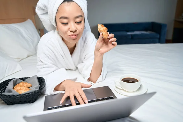 ベッドの上のラップトップを使用して白いコートのアジアの女性 女性はホテルのベッドでコーヒーとクロワッサンを食べる — ストック写真