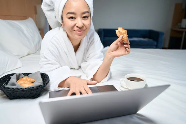 彼女の頭にタオルでアジアの女性の作物の写真 ノートパソコンを使いホテルの部屋でクロワッサンを食べる女 — ストック写真