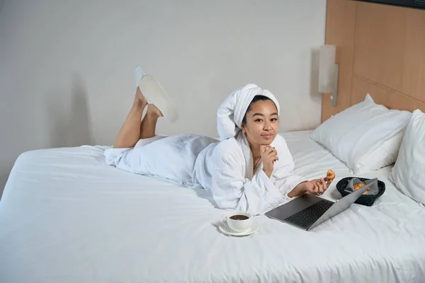 ラップトップを使ったバスローブの女性はホテルのベッドで朝食を取っています — ストック写真