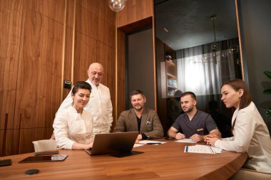 Bir grup doktor masada oturuyor ve gülümseyen bir kadının ofiste dizüstü bilgisayarla çalışmasını izliyorlar.