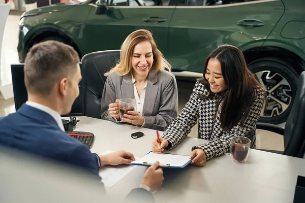 Charming asian woman signs a contract for the purchase of a car, her friend and a car dealership manager are nearby