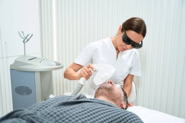 Koruyucu gözlüklü bir güzellik uzmanı orta yaşlı bir adam için fotorejuvenasyon prosedürü uyguluyor. Bir uzman modern ekipman kullanıyor.