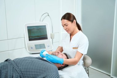 Güzel doktor orta yaşlı bir adama kitle kaldırma ameliyatı yapıyor. Hasta kozmetoloji kanepesinde yatıyor.