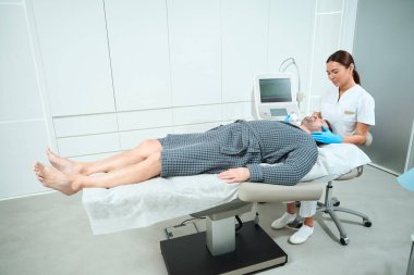 Güzel bir kozmetik uzmanı orta yaşlı bir adama kitle kaldırma ameliyatı yapıyor. Hasta kozmetoloji kanepesinde yatıyor.