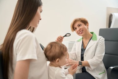 Elinde büyüteçle gülümseyen pediatrik göz doktoru elinde bebekle bir kadının önünde sandalyede oturuyor.