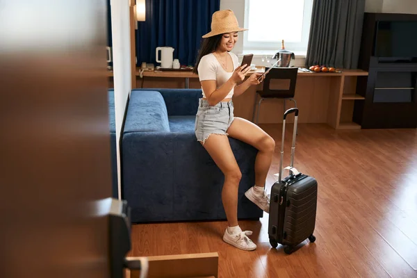 Gülümseyen Kadın Turist Koltukta Oturuyor Tekerlekli Bavulun Yanında Pansiyondaki Telefonu — Stok fotoğraf