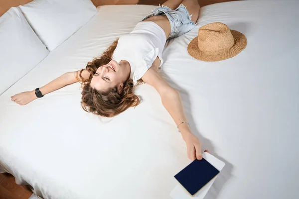 快乐的女性游客躺在靠近帽子的床上 在酒店房间里持票 — 图库照片