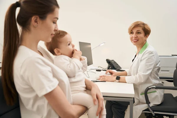 Uśmiechnięty Lekarz Pediatra Siedzący Przy Komputerze Patrzący Młodą Kobietę Dzieckiem — Zdjęcie stockowe