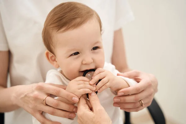 Little baby gnawing at magnifying glass in ophthalmologist hand in mother presence