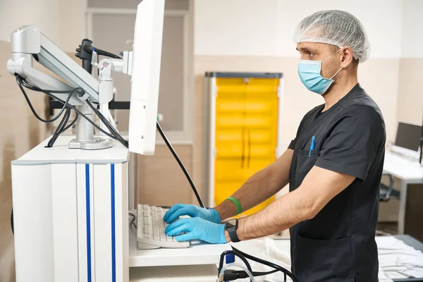 Tıbbi Maskeli Ciddi Bir Teşhisci Bilgisayar Klavyesinde Monitör Ekranına Bakarken — Stok fotoğraf