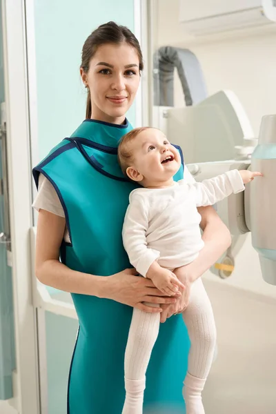 stock image Pleased woman in lead apron holding smiling little child in her arms in front of x-ray machine