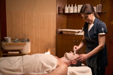 Gülümseyen kozmetik uzmanı hasta cildinde maske takıyor.