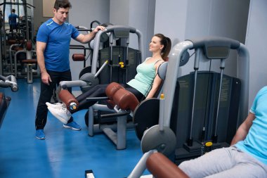 Neşeli, olgun bir kadın spor aletlerinin üzerine oturmuş spor hocasının rehberliğinde bacak uzatıyordu.