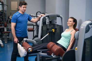 Gülümseyen sporcu kadın kişisel antrenörünün rehberliğinde spor aletlerine bacağını uzatıyor.