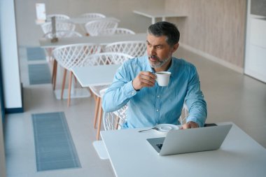 Yetişkin bir adam masada oturmuş çay içiyor, yemek odasında dizüstü bilgisayarda yazıyor.