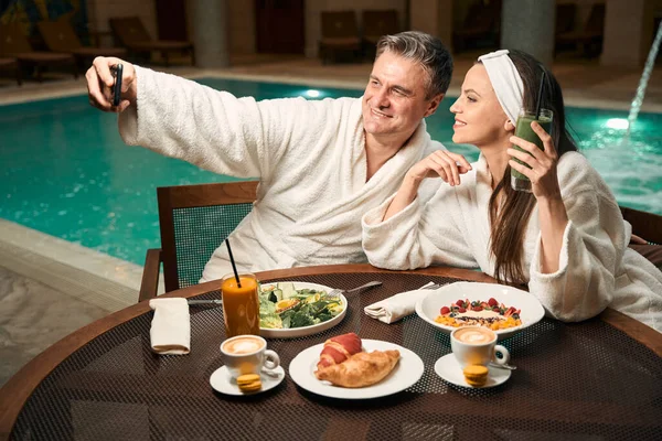 在游泳池边的餐桌前 笑着的男人和他快乐的女伴摆出一副智能手机相机的姿势 — 图库照片