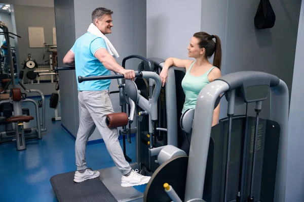 stock image Pleased sporty man performing side leg raise on exercise machine in presence of smiling fitness partner