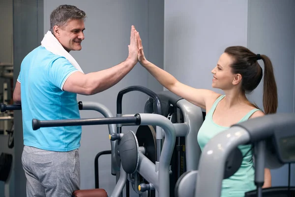 Smiling sporty lady seated on exercise machine giving high five to her joyous fitness partner at gym