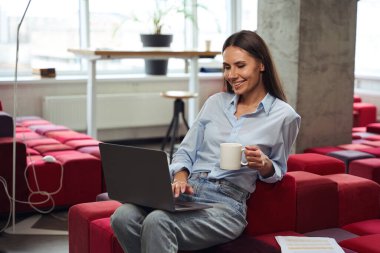 Cheerful young woman with teacup in hand looking at computer monitor while sitting on chair in comfortable coworking space clipart