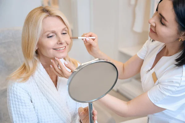 美容師の女性は鏡を見て座っているクライアントのための医療手順を実行します 美容師の予約で女性 — ストック写真