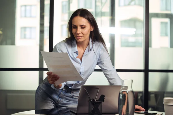 Focused corporate worker leaning on desk before laptop while looking at document in her hand