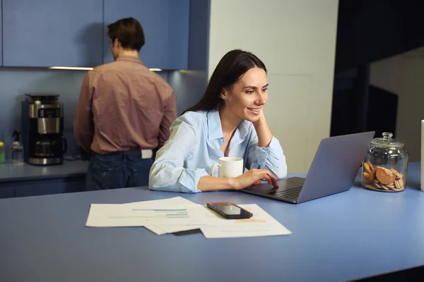 一位快乐的女员工坐在桌旁 一边在笔记本电脑屏幕上看书 一边为自己煮咖啡 — 图库照片