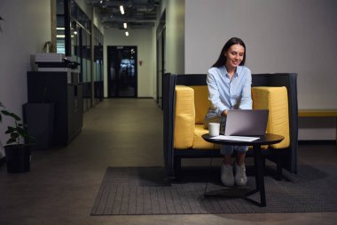 Gülümseyen şirket çalışanı dinlenme odasındaki masada otururken dizüstü bilgisayarda klavye kullanıyor.