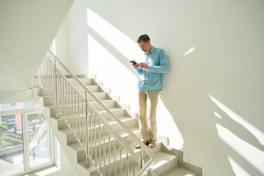 Adam, parlak bir kliniğin koridorunda merdivenlerde duruyor ve telefona bakıyor.