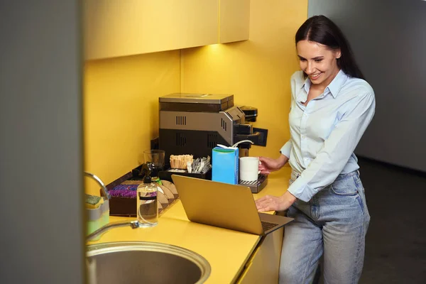コーヒーマシンのドリップトレイにマグカップを配置しながら ラップトップ画面を見て楽しい女性のオフィスワーカー — ストック写真