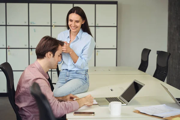彼女の忙しい同僚を見ながらオフィスの机の上に座っているカップと陽気な企業の労働者 — ストック写真