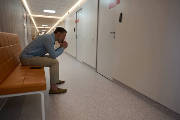 在一家现代化医院的大厅里 一个多愁善感的成年人靠着双手坐在一个褐色的沙发上 旁边有一部电话 侧视图 — 图库照片