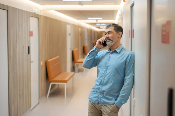 壁にもたれかかる灰色の髭の男が病院の廊下で電話で話している ウエストハイ — ストック写真
