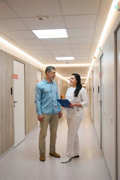Man patient talking with a woman doctor in a white suit in the corridor of a bright clinic