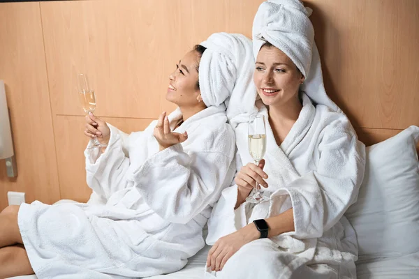 穿着浴衣的女朋友在酒店里喝香槟两个穿着白衣 戴着眼镜的女人在床上 — 图库照片