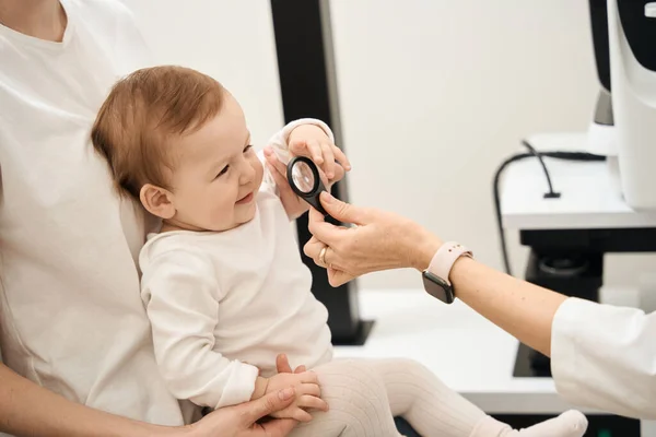 女眼科医生手拿着放大镜 坐在坐在母亲膝上笑着的婴儿面前 — 图库照片