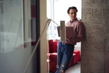 Modern ofisteki sütuna yaslanırken dizüstü bilgisayarı elinde tutan düşünceli genç bir şirket çalışanı.