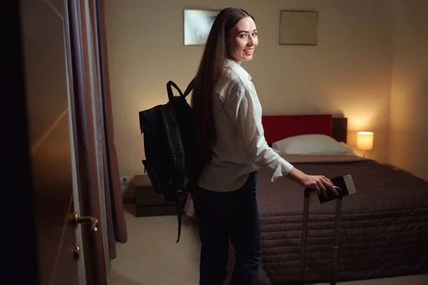 大きなベッドの近くのホテルのベッドルームには バックパックとスーツケースを着た若い女性が立っています バックビュー — ストック写真
