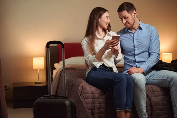 ホテルの部屋の大きなベッドに座っている間 若い男と女は一緒に電話を見ています — ストック写真