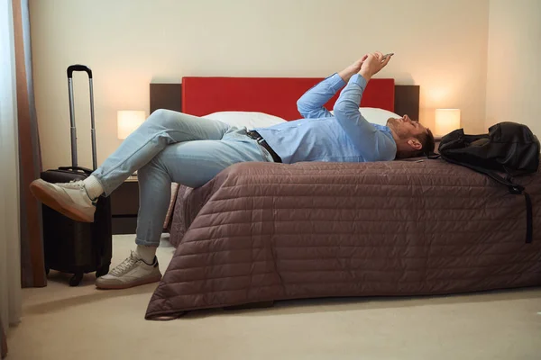 Красивый Парень Просматривает Телефон Лежа Большой Кровати Чемоданом Рядом Ним — стоковое фото