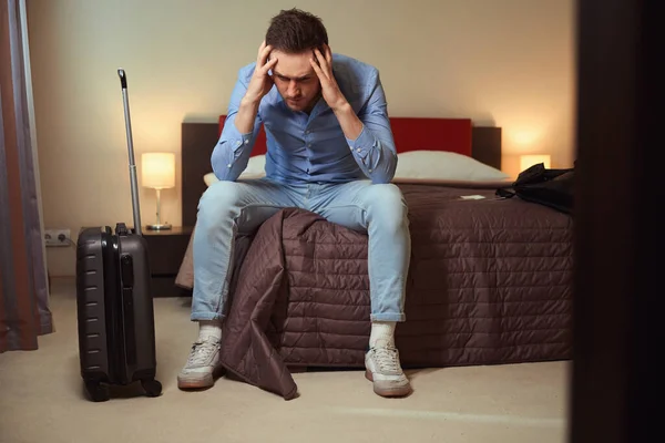 在一个现代化的酒店房间里 一个头疼的可悲的家伙坐在一个大床上 旁边有一个手提箱 — 图库照片