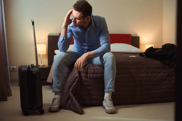 在一个现代化的酒店房间里 一个头痛的可悲的男人坐在一个大床上 旁边有一个手提箱 — 图库照片