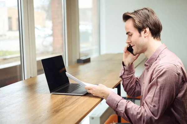 スマートフォンで話している間にノートパソコンを見て文書を見て座っている笑顔のオフィスの従業員の側面図 — ストック写真