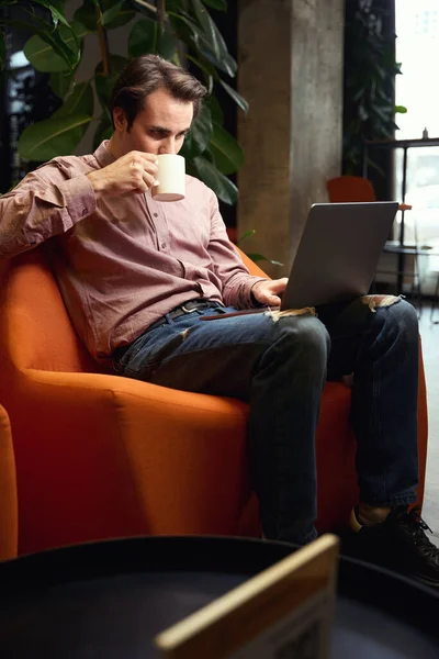 同僚のスペースでポータブルコンピュータに取り組んでいる間 若い男は熱い飲み物を飲む — ストック写真