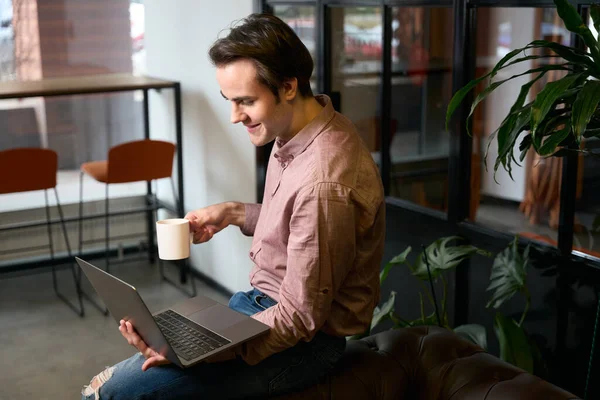 Pleased guy with portable computer and mug in hands sitting on sofa arm in coworking space