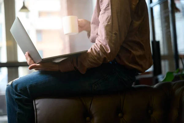 Cropped photo of man with laptop and mug in hands sitting on sofa back in coworking space