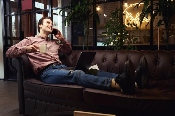 陽気な男座っています上の居心地の良いソファとノートパソコンとマグカップの熱い飲み物の電話会話中にコワーキングスペース — ストック写真