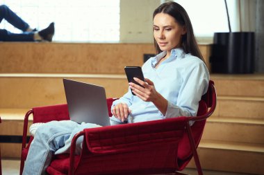 Dizüstü bilgisayarı ve akıllı telefonu olan odaklanmış genç bir kadın dinlenme odasındaki koltukta oturuyor.