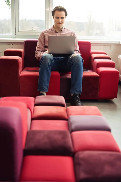 会社のオフィスで彼のラップトップを使用してソファの上に座って穏やかな焦点を当てた若い企業の労働者 — ストック写真