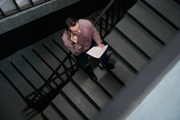 Top view of corporate worker with pile of documents standing on stairs during smartphone conversation