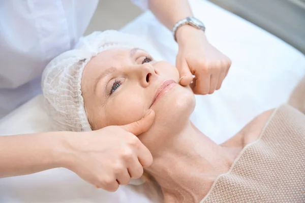 Happy adult lady in cosmetic cap getting facial massage and stroking the chin